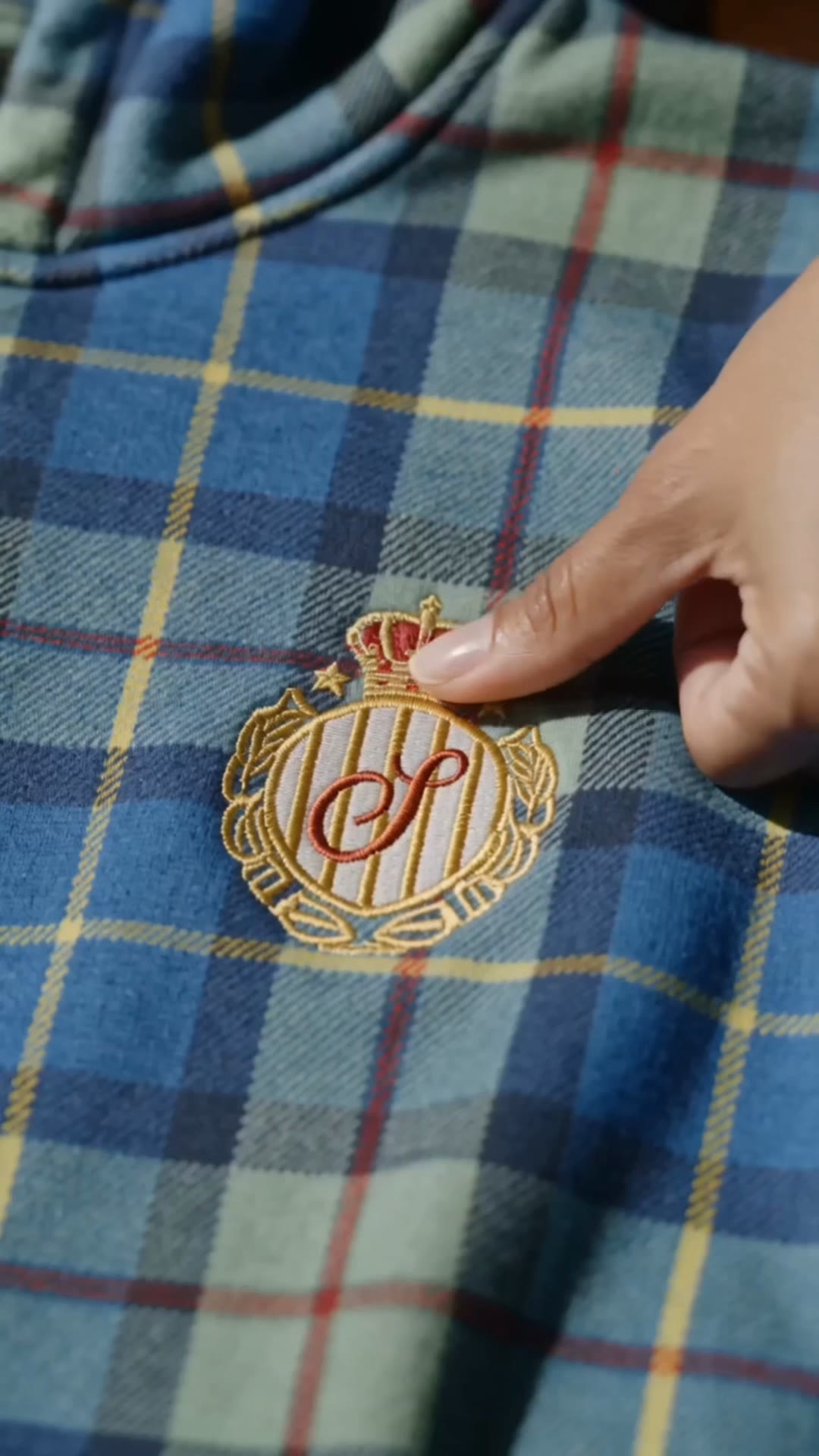 Macro frame from film three: a thumb sweeping the gold laurel wreath edge of the Shinzo chest crest on turquoise plaid
