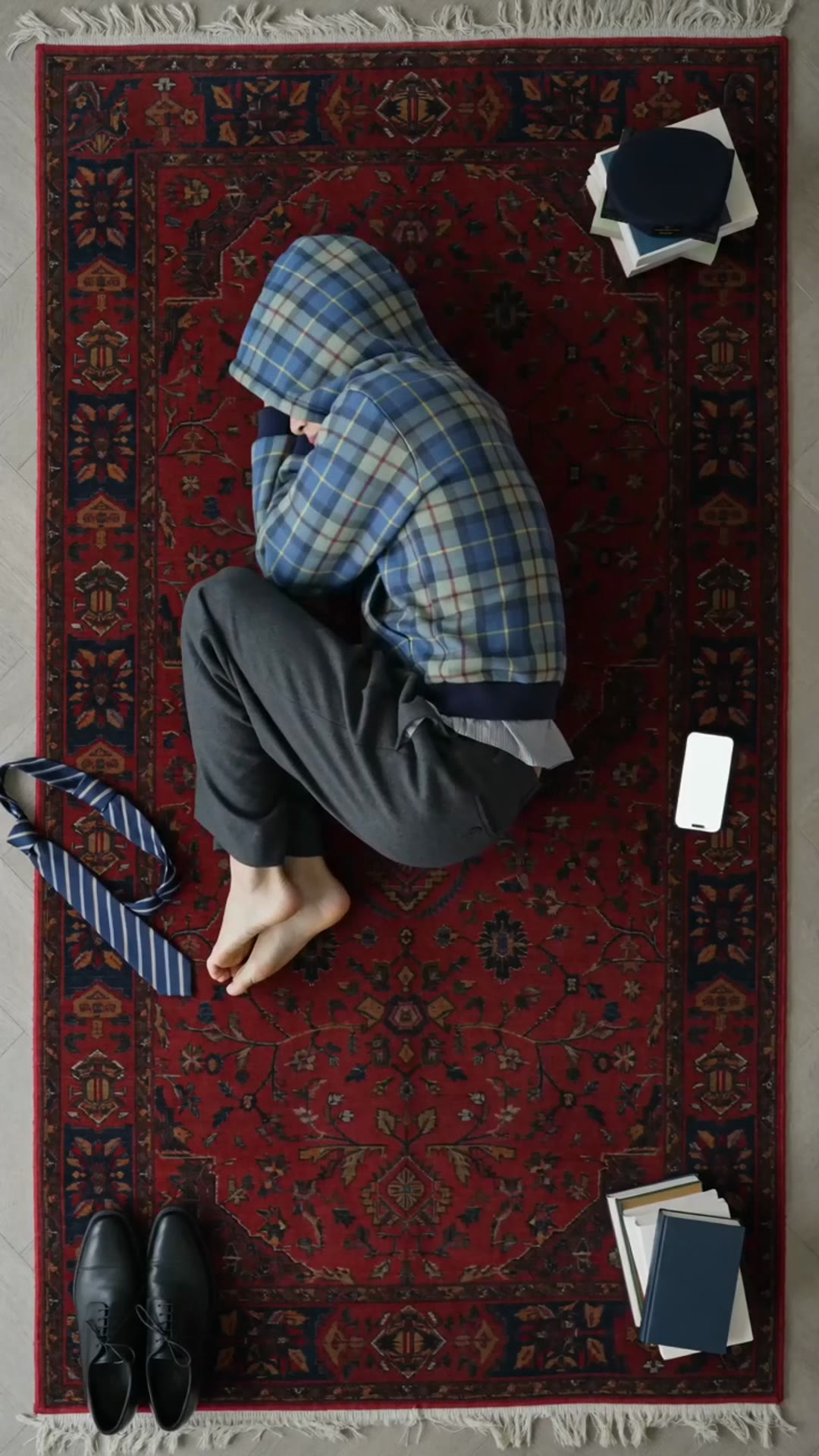 Locked overhead frame from film four: the wearer curled on a deep red Persian rug with the hood up, phone glowing beside him
